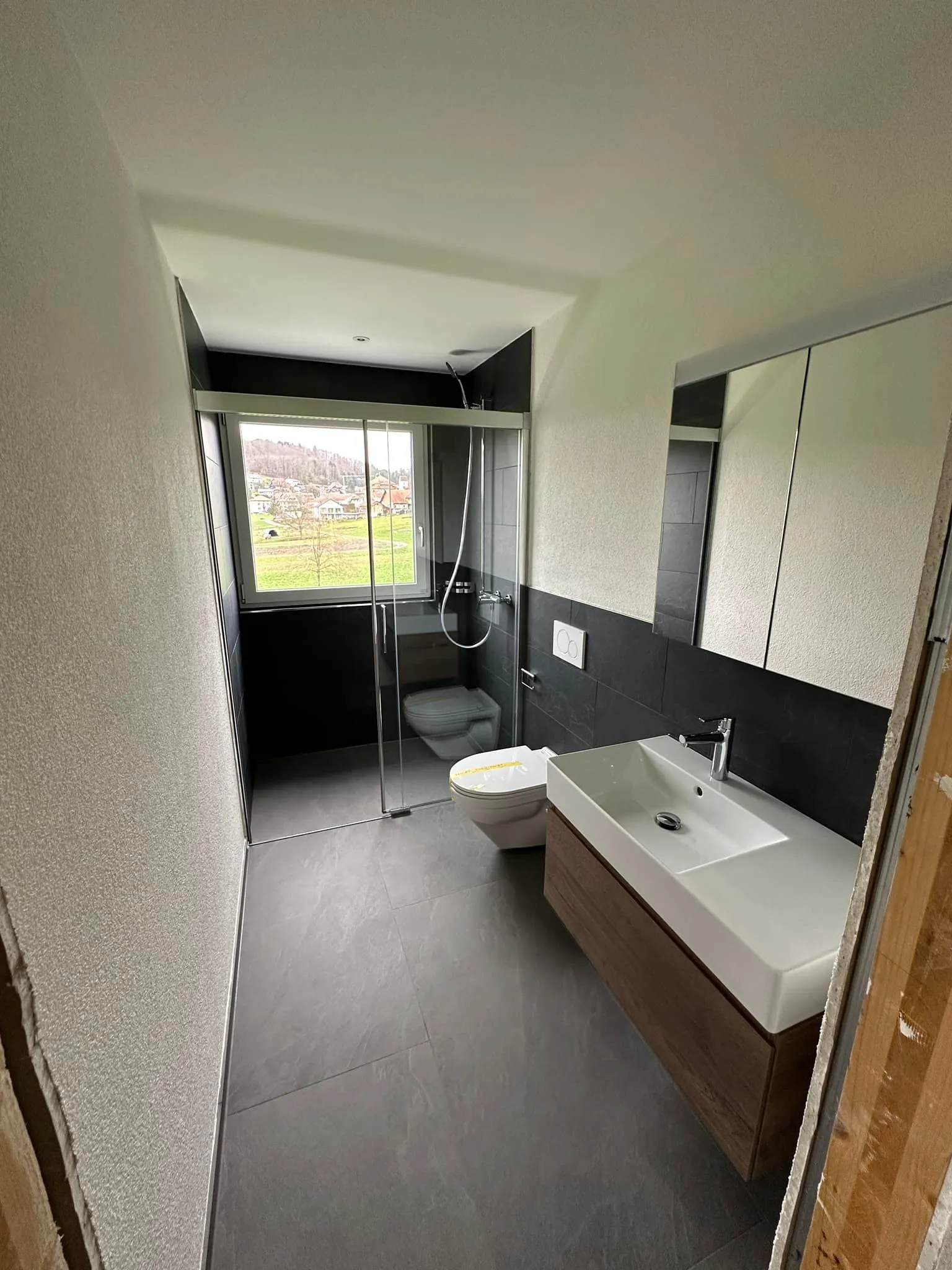 Duschbad mit Glastrennwand, dunklen Wandplatten und grossformatigem Boden.