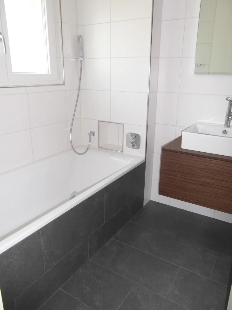 Badezimmer mit Badewanne, weissen Wandplatten und dunklem Bodenbelag.