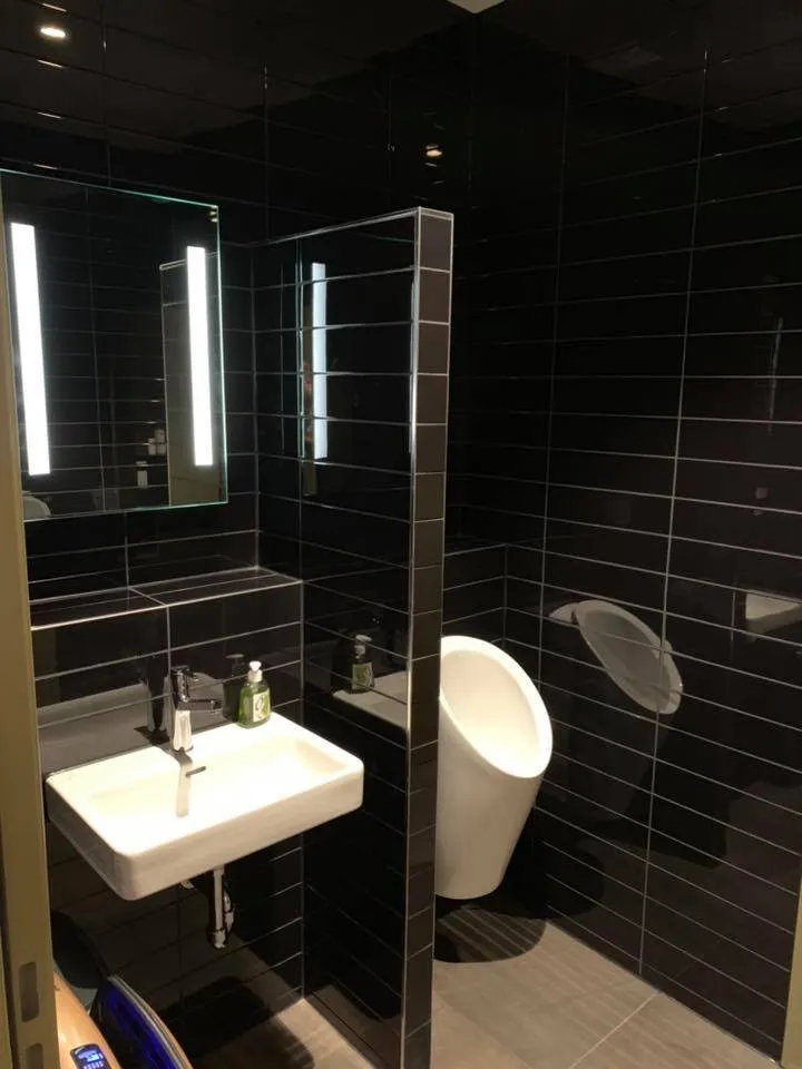 Herren-WC mit dunklen Wandplatten, Urinal und beleuchtetem Spiegel.