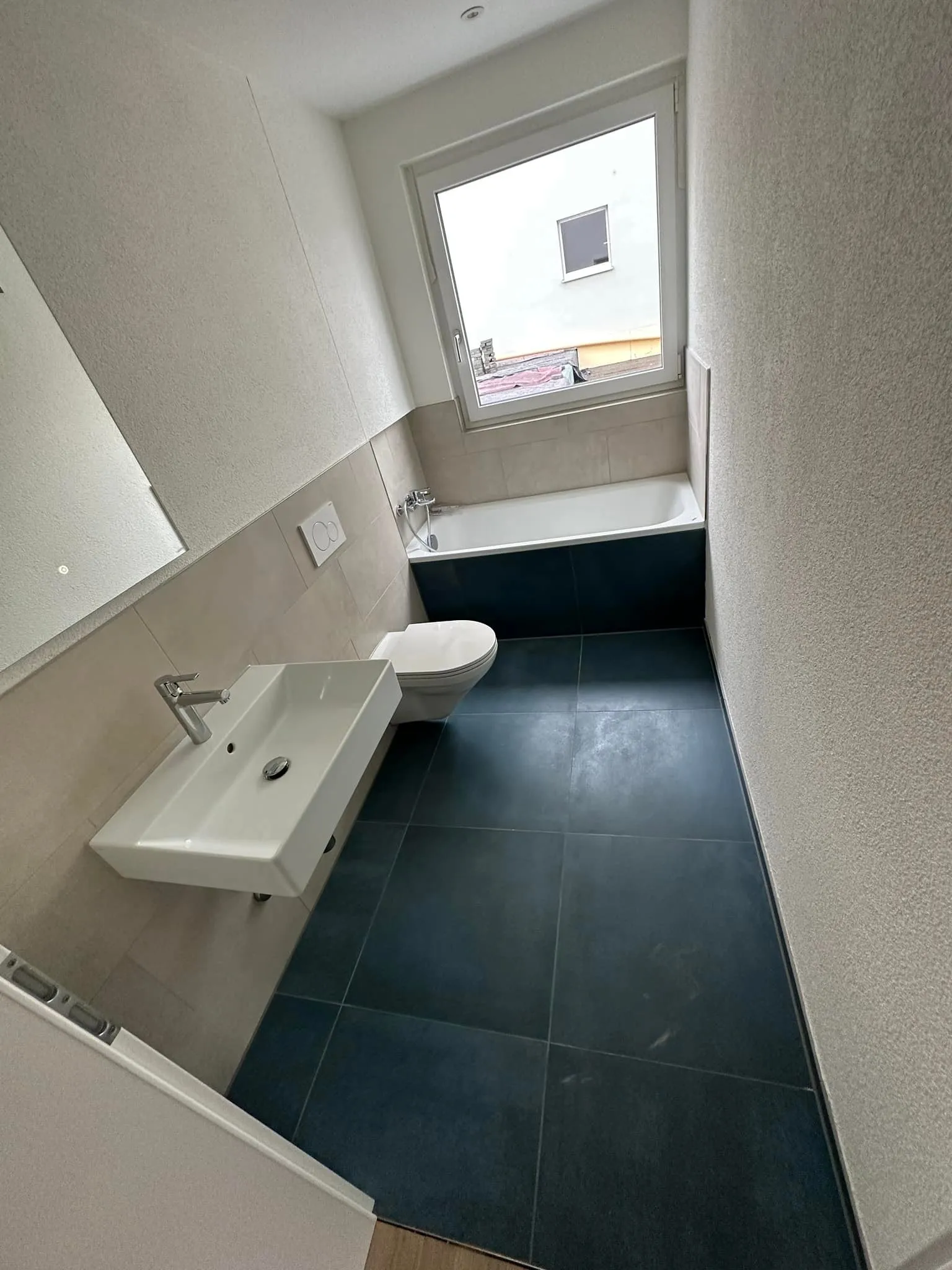 Badezimmer mit Wanne unter dem Fenster und dunklen Bodenplatten.