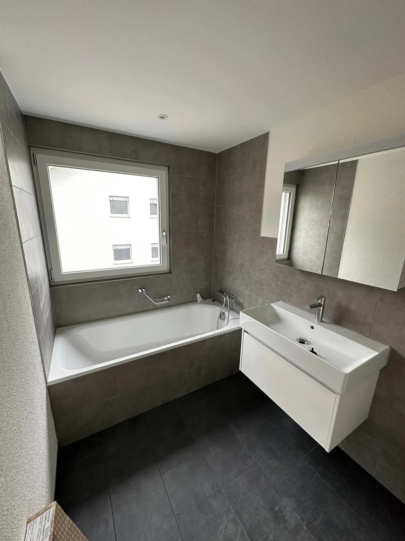 Badezimmer mit Wanne, grauen Wandplatten und weissem Waschtisch.