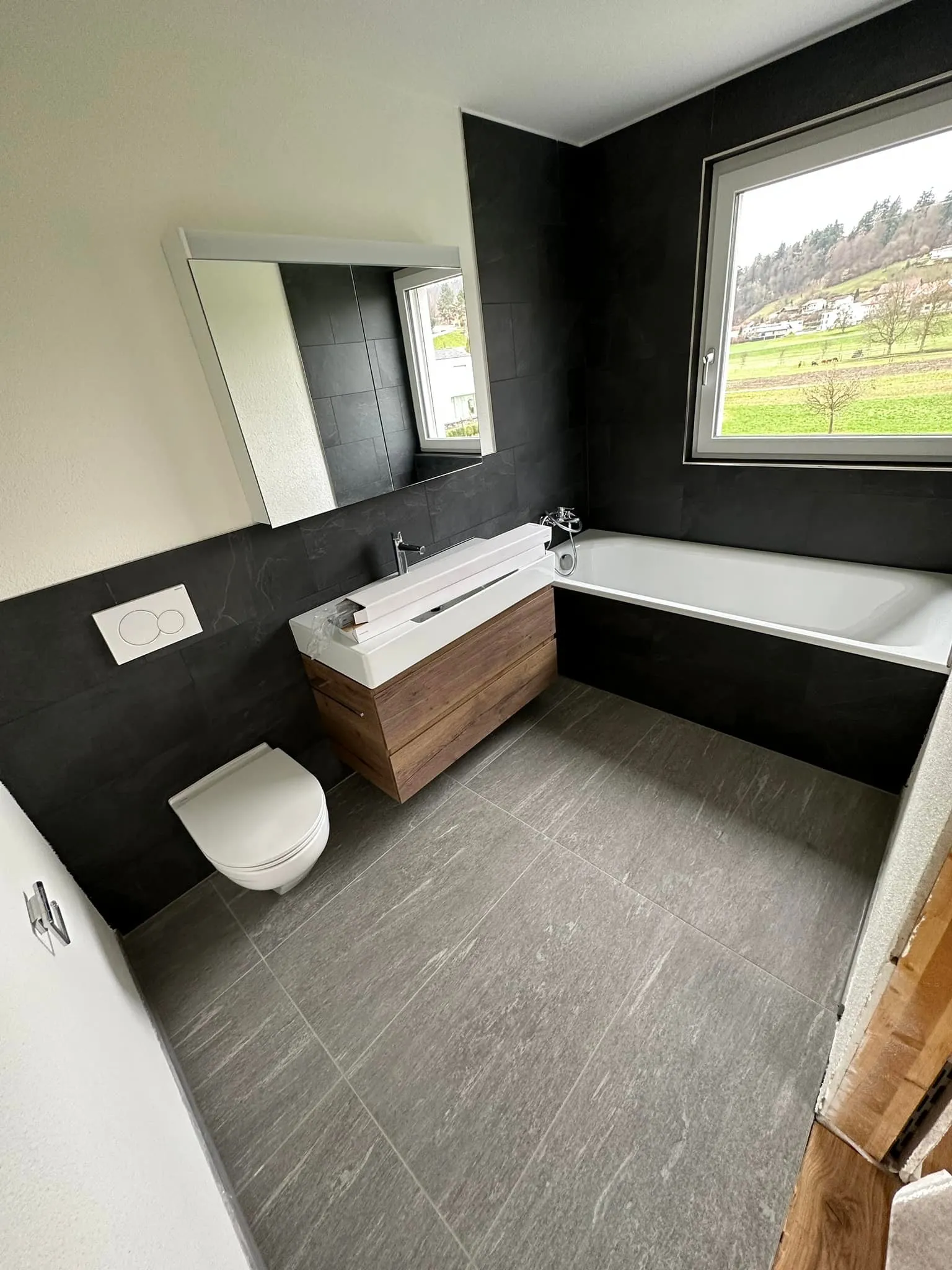 Badezimmer mit dunkler Plattenverkleidung, Wanne und Ausblick ins Grüne.